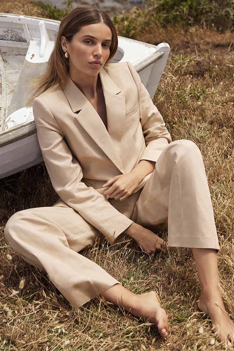 a model with long straight highlighted hair wears a camel coloured suit, she sits on a sunbleached grass atop a cliff, she leans her back against an old whitewashed boat 