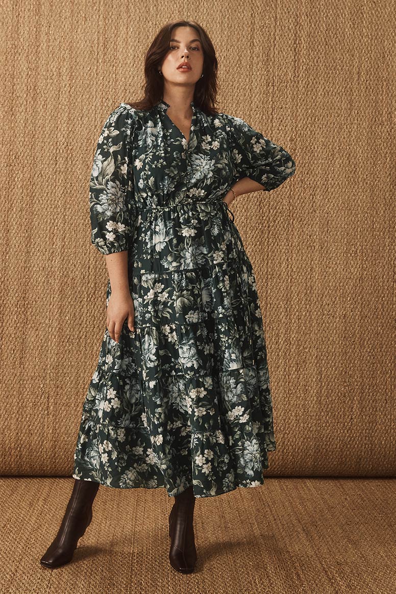 curvy model with dark hair wearing a long sleeve floaty dress in a deep green floral print with sleek choc brown boots, she poses with one hand on her hip, the floor and the wall looks to be covered in gold hessian material