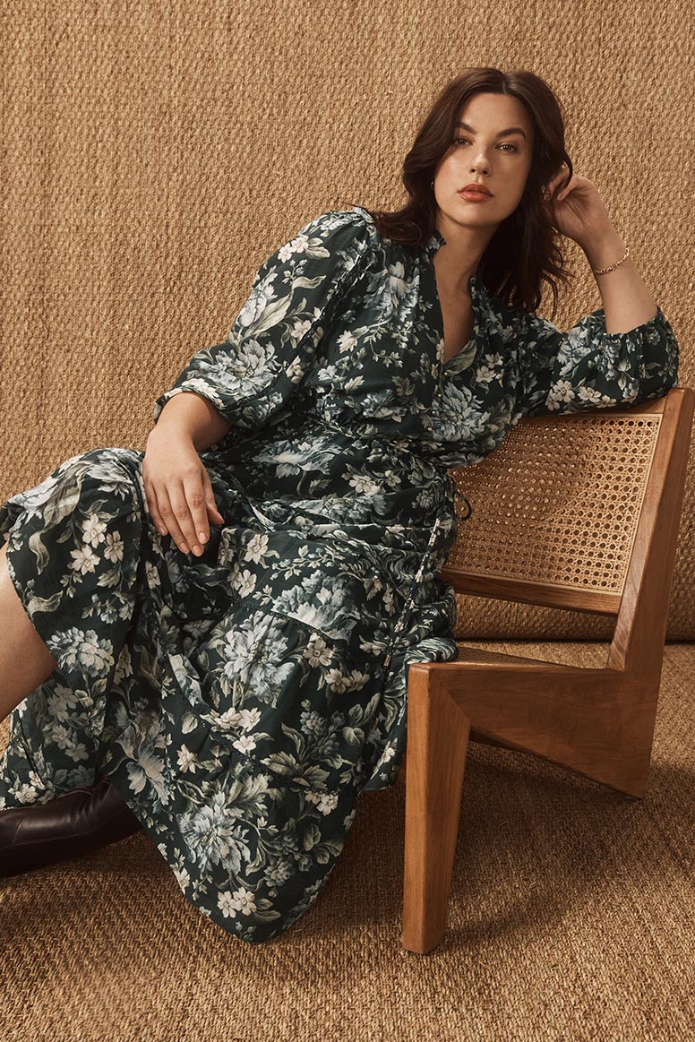 curvy model with dark hair sits on a rattan low chair wearing a dark green floral dress, the wall behind her is covered with a warm golden hessian material