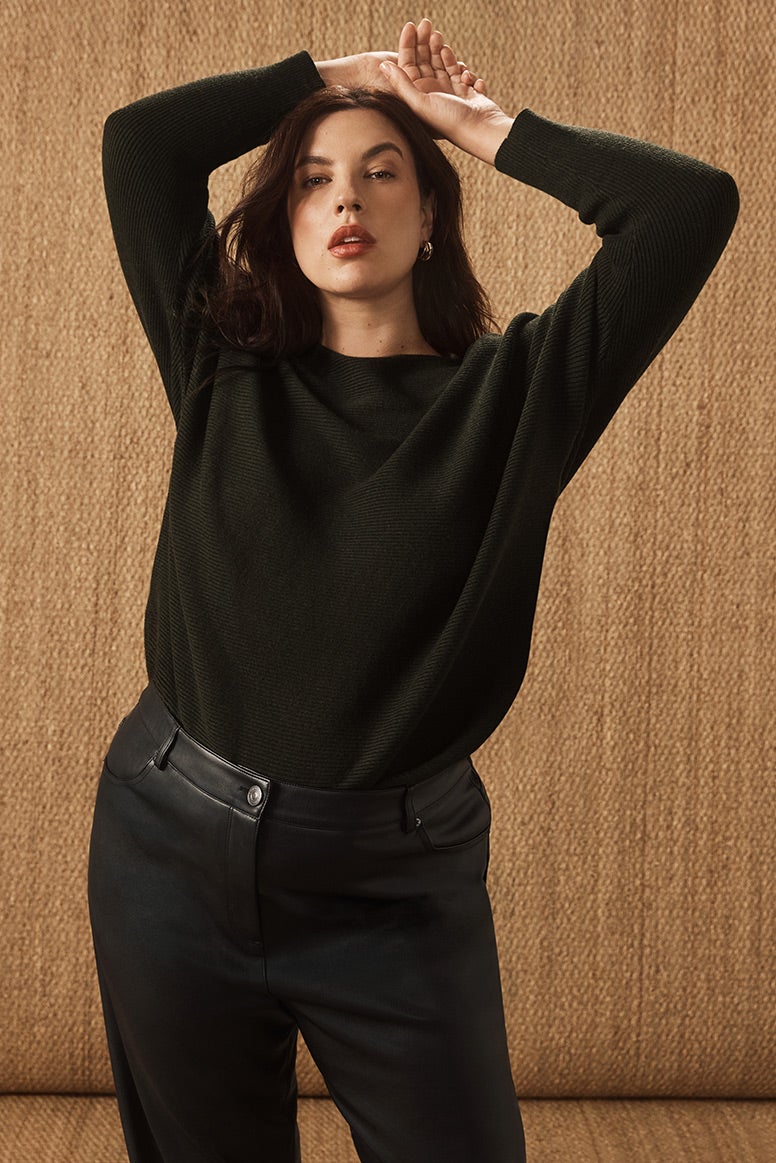 curvy model with dark hair wearing a black knit top tucked into black leather pants, she poses with her hands on her head, the floor and wall are covered in a golden natural hessian material