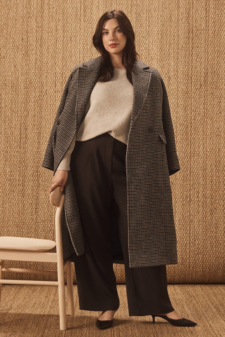 curvy model with dark hair poses standing up resting one hand on a back of chair just off screen, she is wearing a camel knit black pants and a checked coat draped over shoulders, she wears heels and the floor and the wall is covered in hessian materials