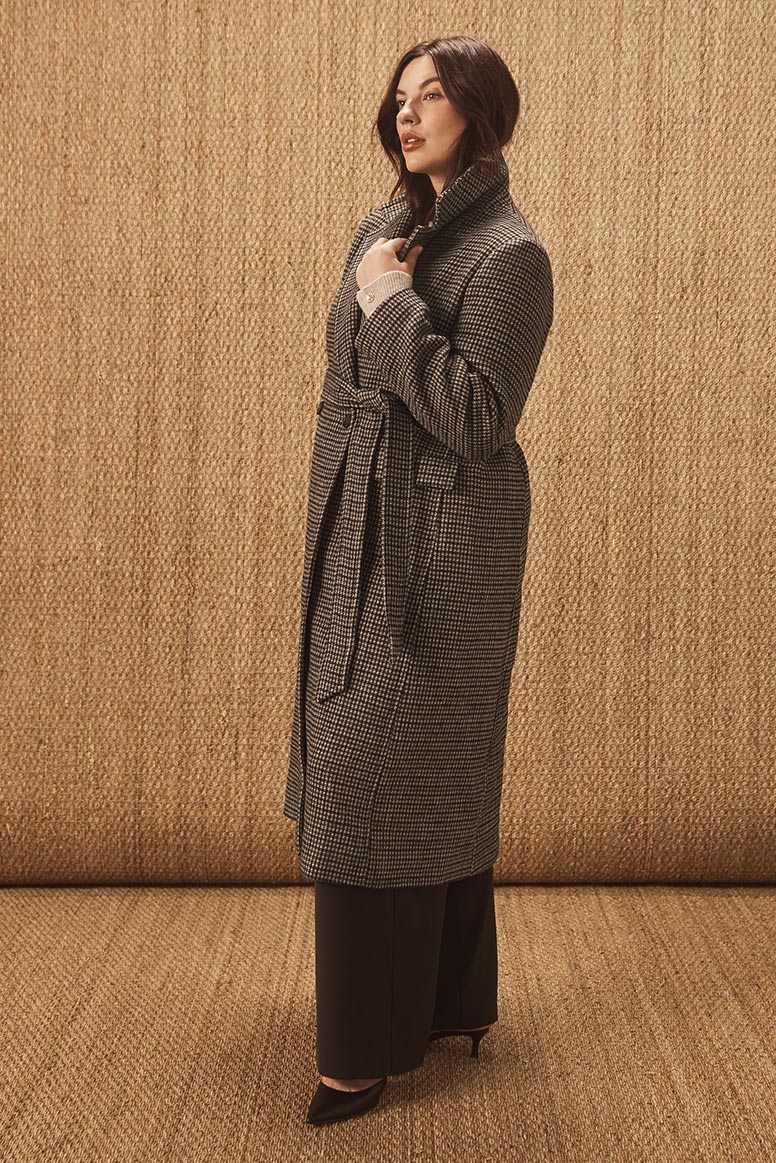 curvy model with dark hair standing in profile wearing a checked coat belted with black pants and matching pointed toe heels, the wall and floor is covered with a golden hessian material