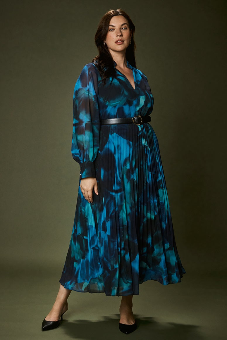 brunette curve model wearing a long sleeve electric deep blue abstract floral print dress with black pointed heels, a moody deep green wall is her background