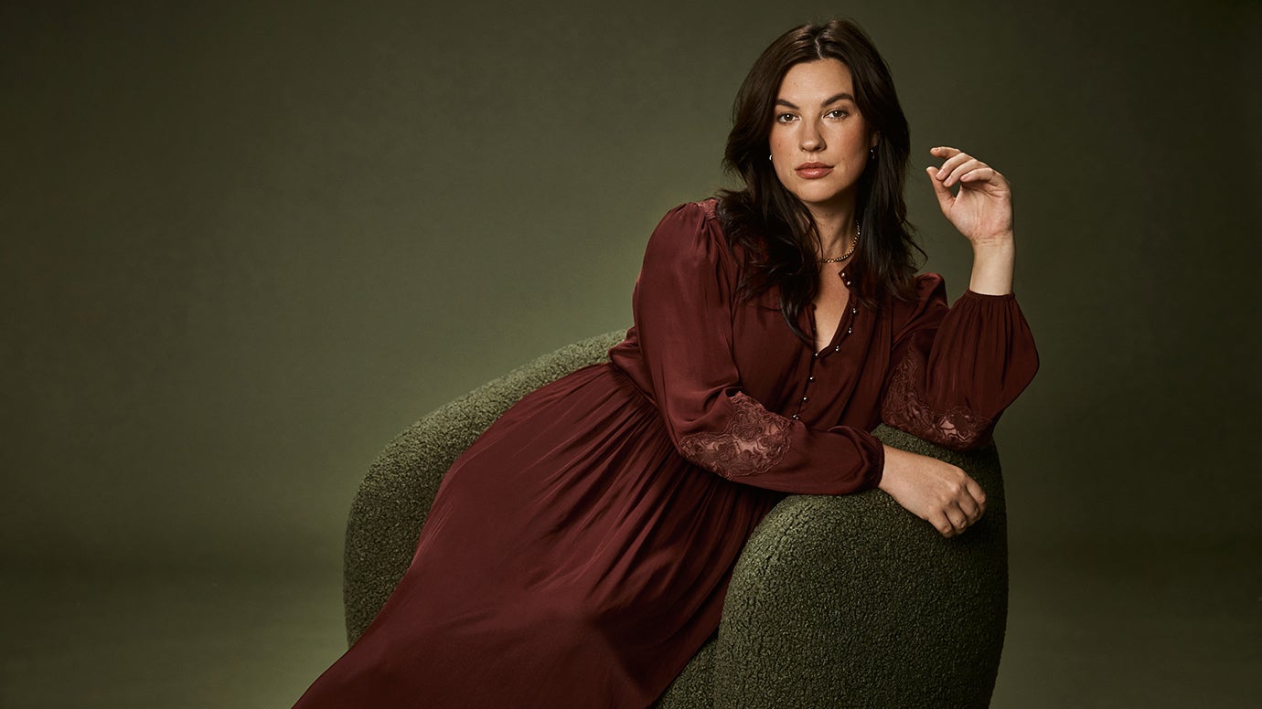 brunette curve model reclining on deep green armchair wearing a deep cherry/lum coloured dress with long sleeves