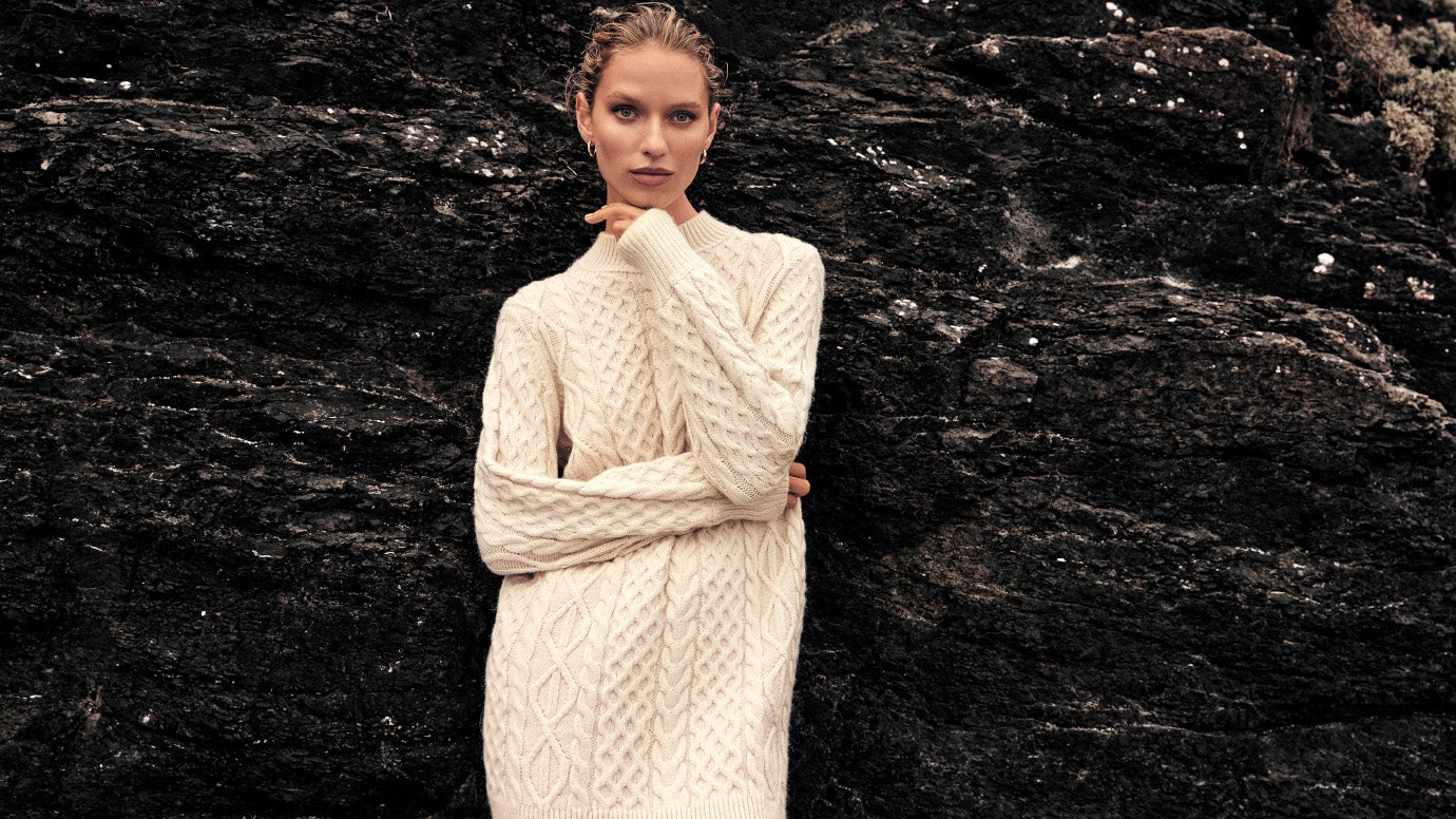 model in chunky cable knit dress in cream standing in front of dramatic black cliff face