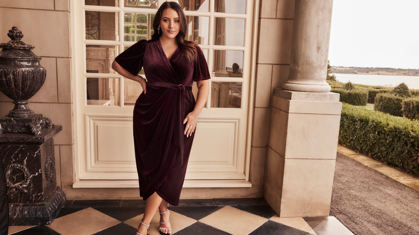 plus size model berry coloured velvet wrap dress standing in front of an elegant villa 