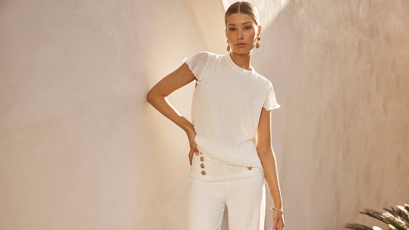 a model poses with one hand on hip against a fancy looking concrete wall wearing a white blouse and white pants with gold buttons, she is bathed in warm sunlight