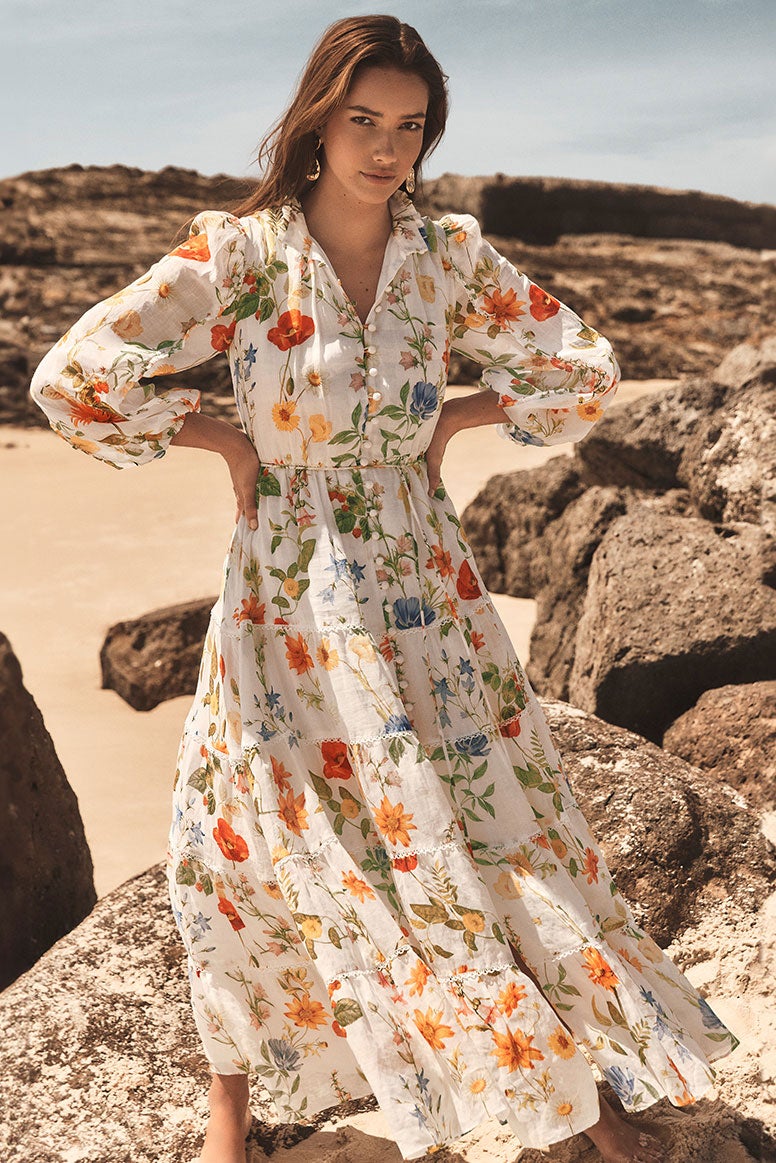 model with long brown hair poses sitting on a rock with sand and the water behind her, she wears a white mini dress with puff sleeves, it has a multi colour floral print