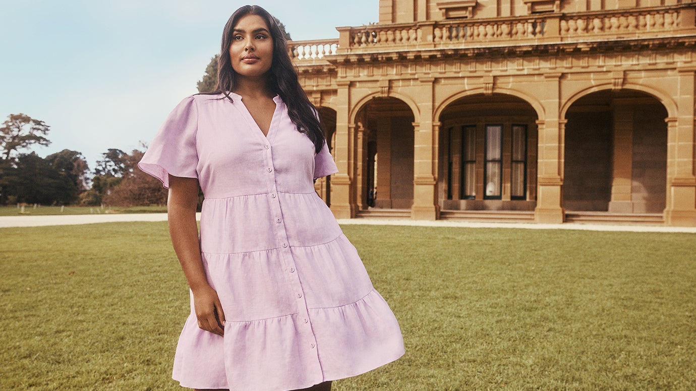 plus size model shanaya peters in a soft pink tiered linen mini dress with short sleeves and buttons up the front posing in front of stately sandstone building and a green lawn