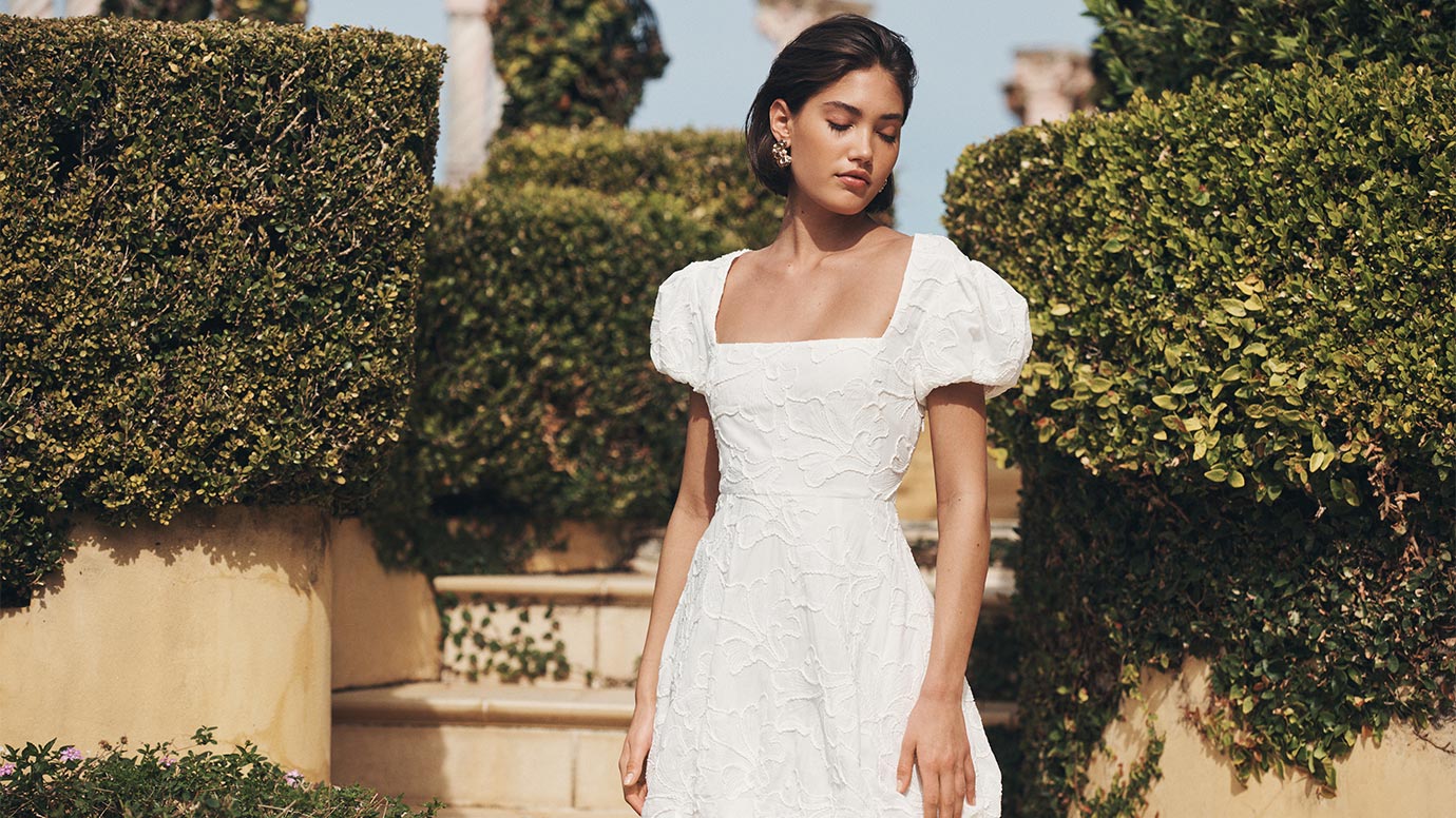 model with a dark brown bob wearing a puff-sleeve white embroidery dress posing in front of mediterranean looking yellow stone steps and large green hedges