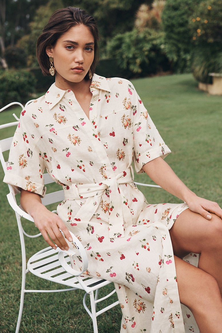 model wearing a cream coloured shirt midi dress with belted waist in a cherry print, sits on a white wire frame lawn chair on a lush green lawn