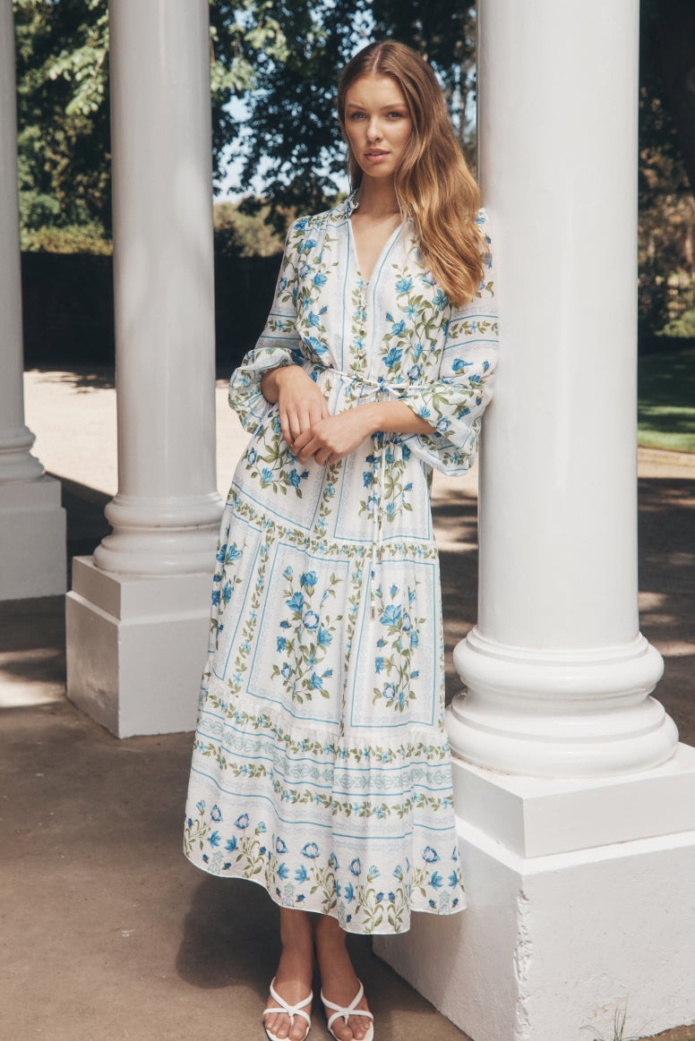 model wear long sleeve flowing white dress with delicate placement print florals in blue and green, she leans against grand white columns