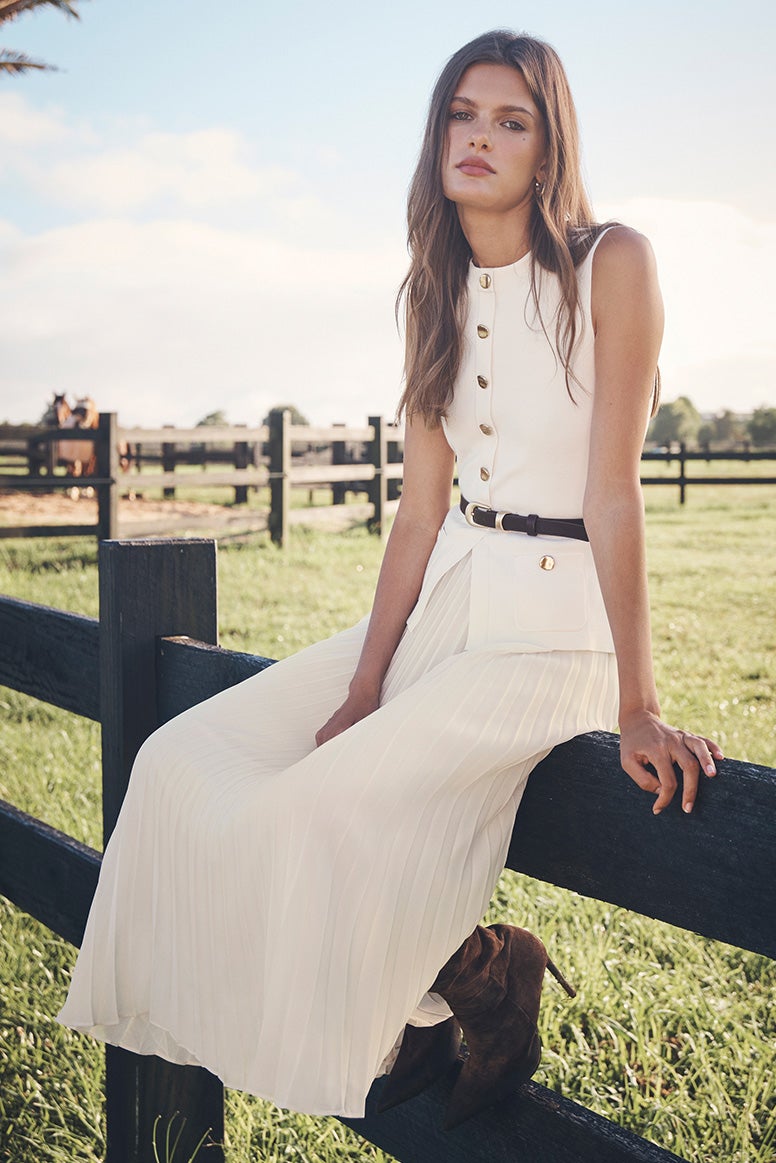 model is standing in profile on a lush green tennis court, she wears a white knit dress with green contrast tipping and sunglasses, white socks and white sneakers, white tennis balls are all around her feet
