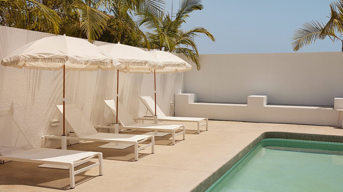 a simple blue pool with concrete rendered white walls enclosing it, palm fronds can be seen just out of frame, white sun loungers and white umbrellas are lined up facing the pool