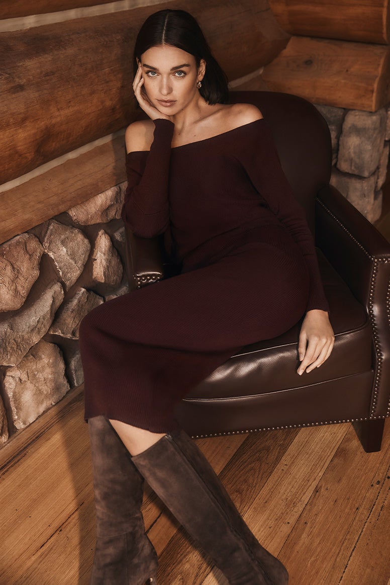 model with brown bob in deep plum knit dress with long sleeves sits on a leather high back armchair in a moodily lit log cabin, she wears knee high brown leather boots