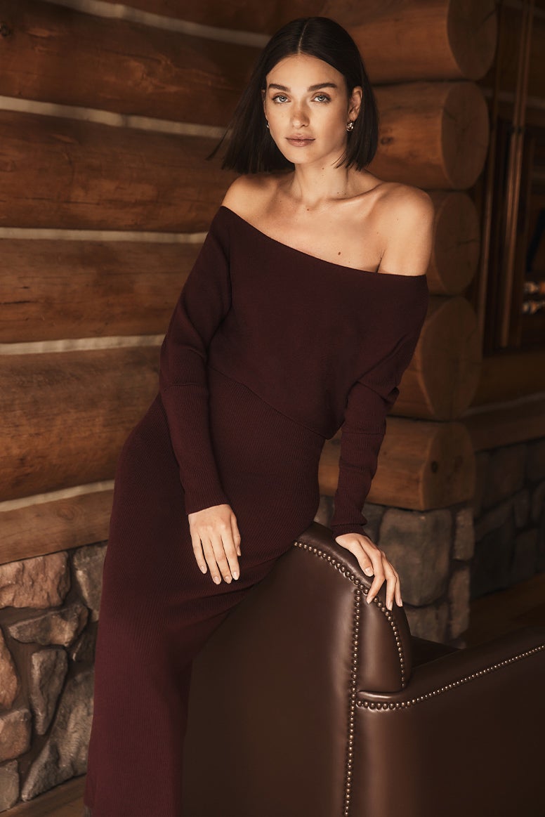 model with brown bob leans against a brown leather armchair in a log cabin wearing an off the shoulder deep plum knit dress 
