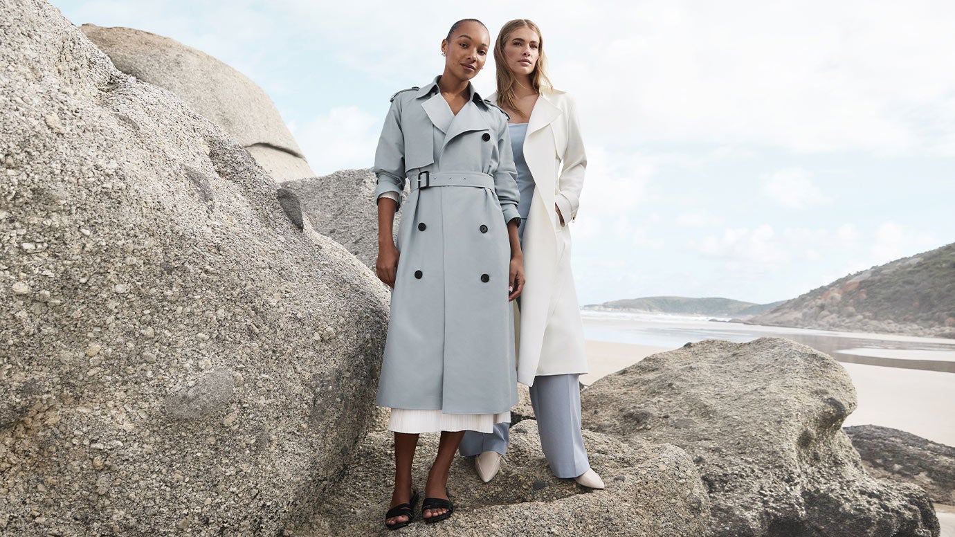 two models posing in front of large grey boulders with the ocean in the background, one is wearing a light powder blue trench, the other is wearing a strapless top and pants in light blue and a cream trench