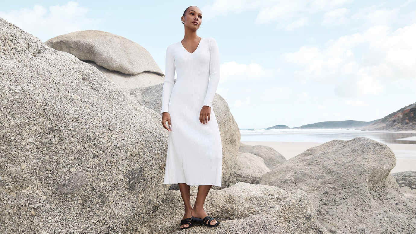 black model with slick back bun wearing a white knit dress with long sleeves and slide sandals, she is standing amongst grey rocks and boulders with the ocean in the background