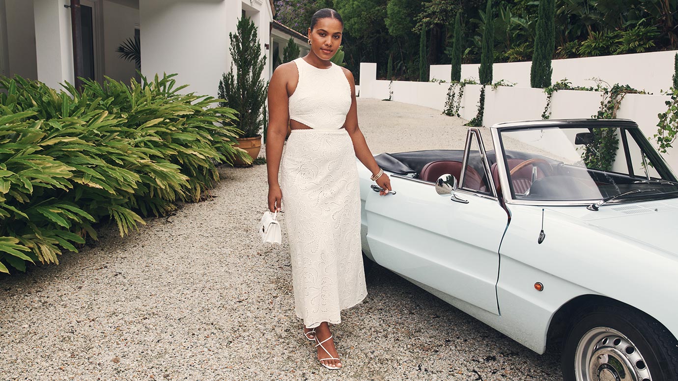 influencer and model sierra johnson wears a white broderie fitted summer dress that has waist cutouts and strappy white heels, she poses in front of vintage convertible car 