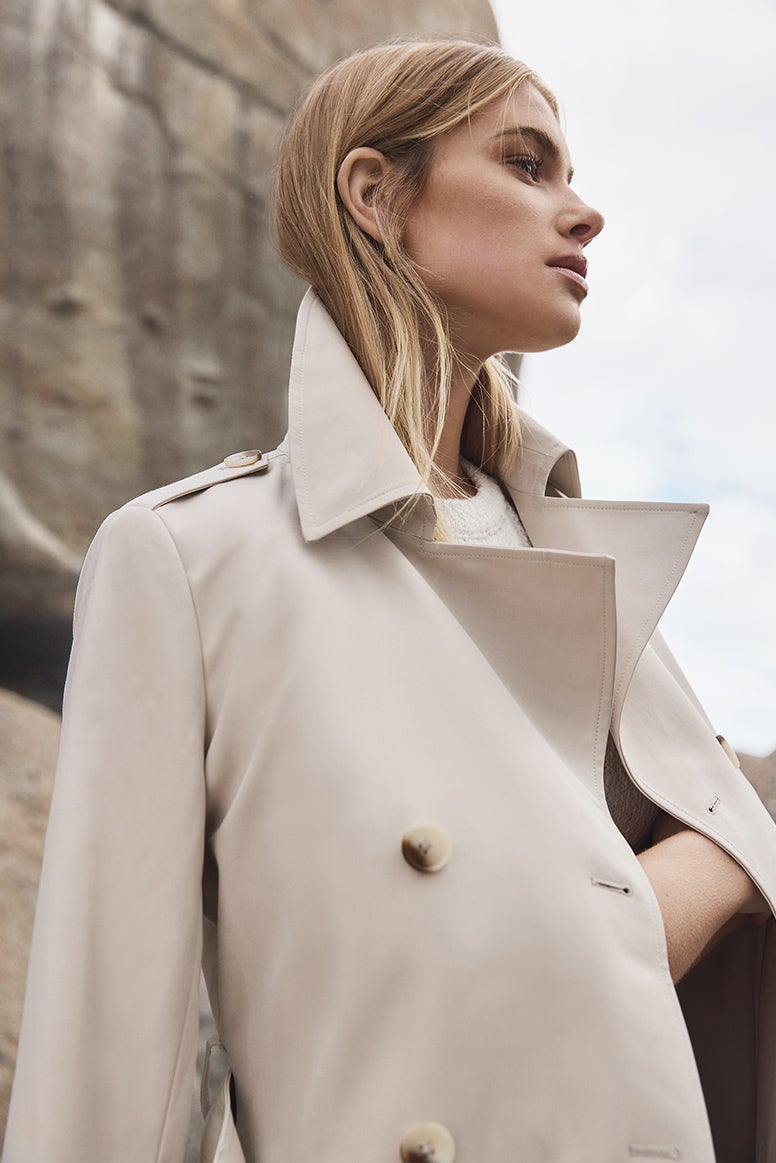 model with sandy coloured straight wearing a sand coloured trench, she is looking off into the distance and has her arms crossed