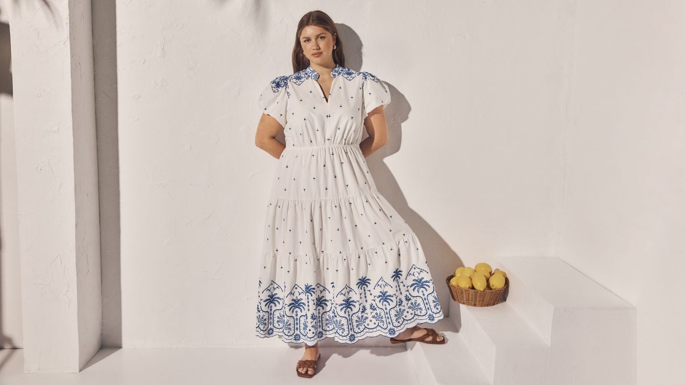 curve model wearing a white tiered maxi dress with blue embroidery standing in front of a white wall with a basket of lemons on a step to her right 