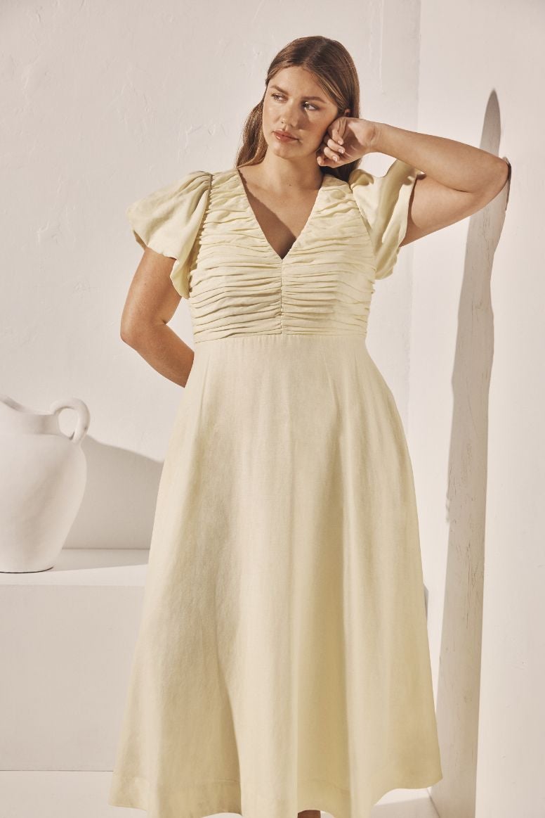 curve model wearing a long flowing lemon hued linen dress leaning against a white wall that looks greek inspired with a white urn in the background
