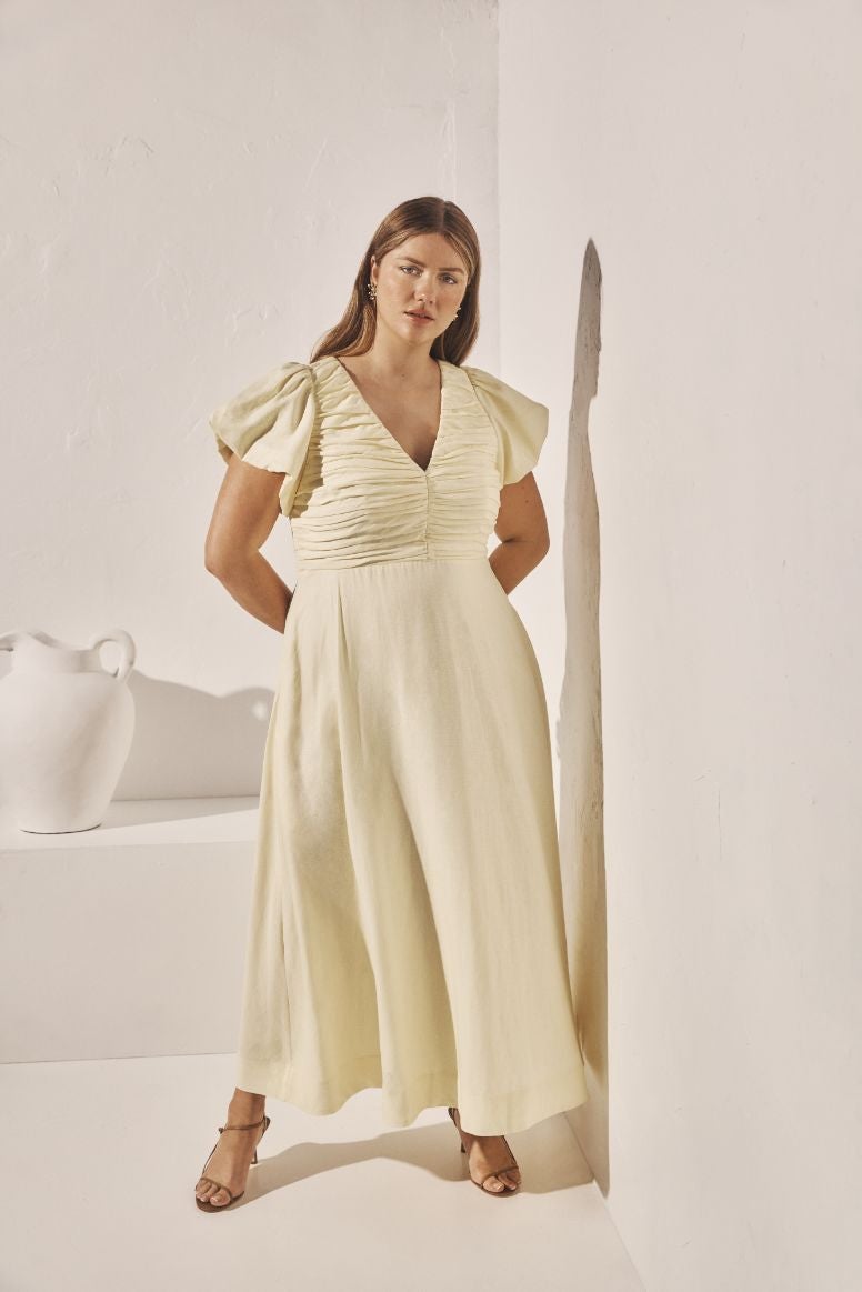 curve model with long hair wearing a lemon coloured linen dress and tan strappy heels, posing in front of white walls with a white urn in the background