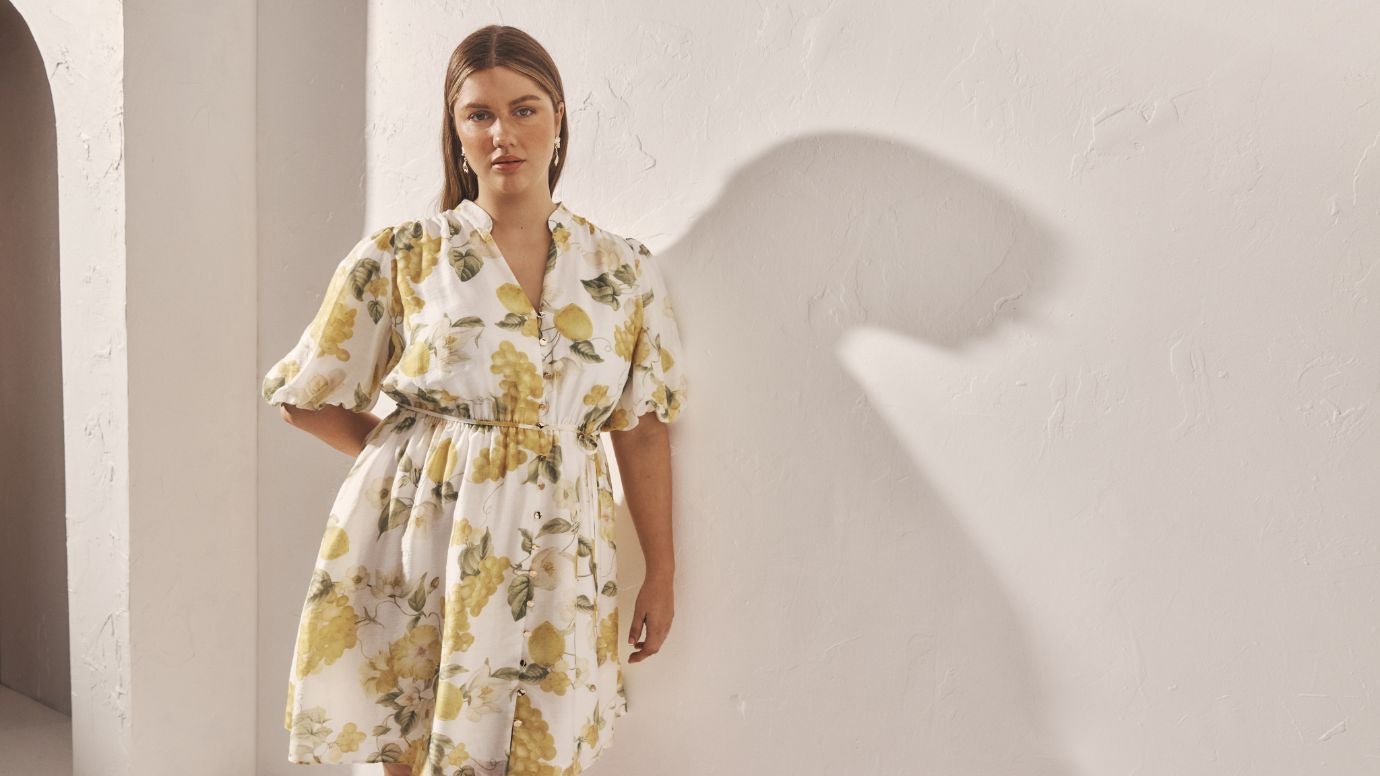 curve model wearing a white and yellow printed dress, the print shows illustrations of grapes, lemon, and florals, she is posing in front of a white wall and her shadow is seen on the wall behind her