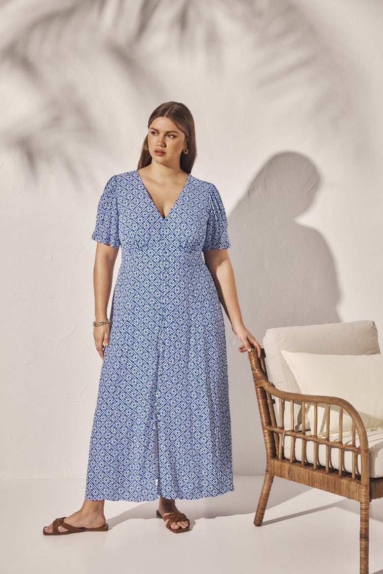 curve model wearing a blue and white tile print sundress with brown sandal slides, she is posing in front of a white wall, her hand is leaning on a rattan chair just out of frame