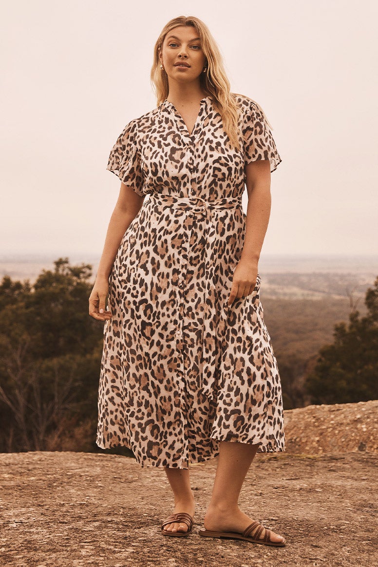 curve model with blonde hair wearing a leopard print wrap midi dress and brown slides, she is posing in a remote bushland landscape