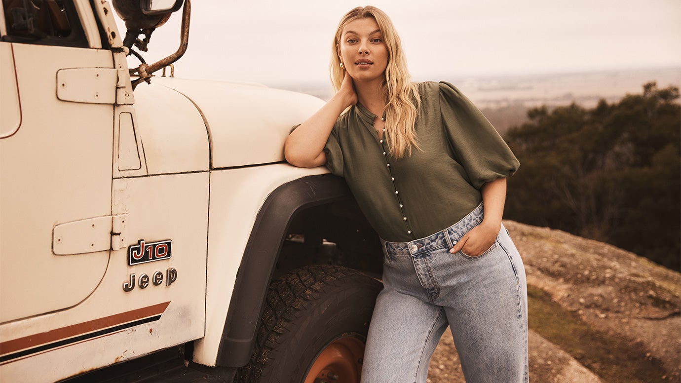 blonde curve model leaning on a vintage jeep parked on a cliff, she is wearing light wash jeans and an army green blouse with flutter sleeves
