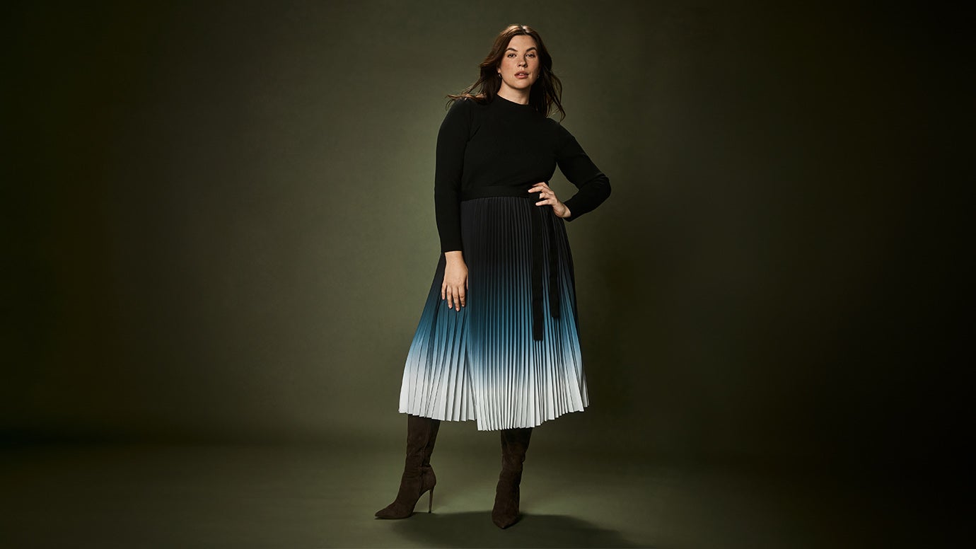brunette curve model stands against a deep green wall and floor, she is standing with one hand on hip wearing a dress that appears like a black knit top and a dip dye pleated skirt with pointed boots, the skirt goes from deep blue to white