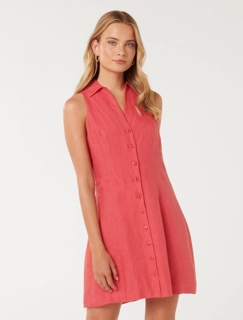 blonde model poses against a white backdrop in a simple sleeveless, linen, mini dress in a watermelon pink colour