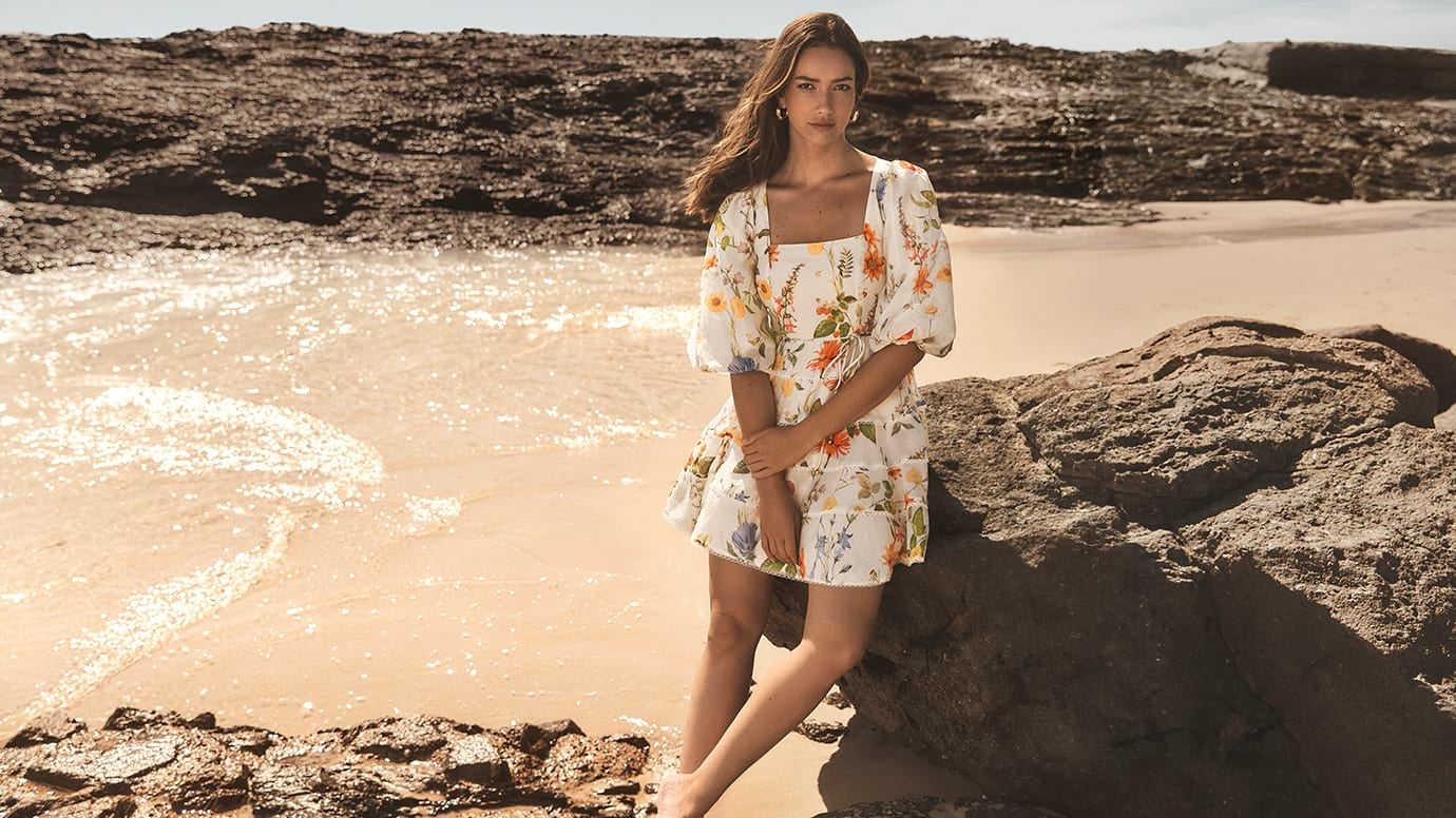 model with long brown hair poses sitting on a rock with sand and the water behind her, she wears a white mini dress with puff sleeves, it has a multi colour floral print