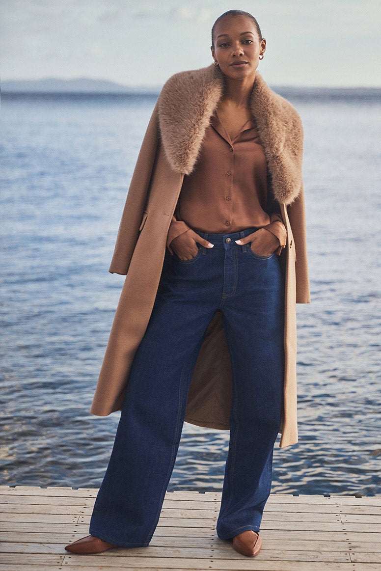 model in a caramel coat with faux fur collar and blue jeans standing on a wharf, sky and blue water is seen in the background