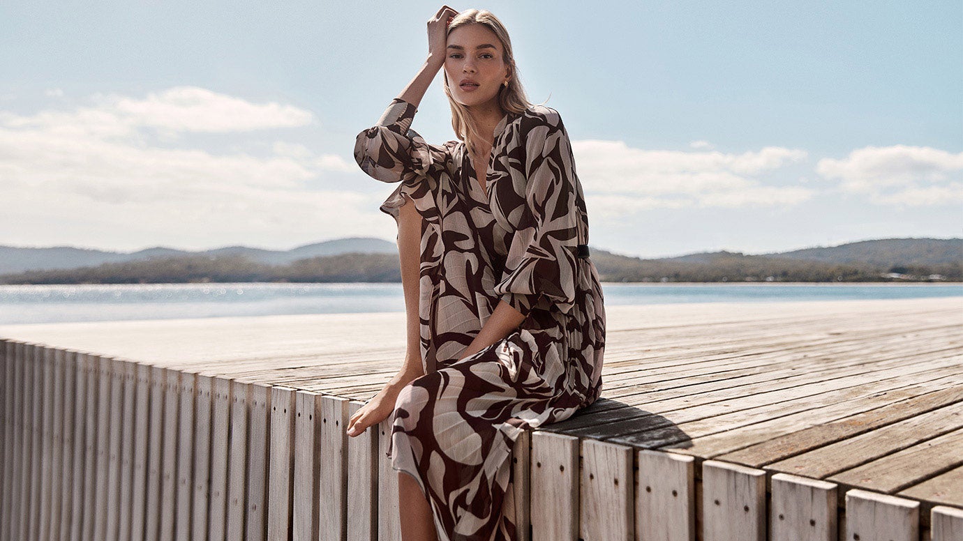 model in a brown and white printed long sleeve dress sitting on a minimal paling style wharf, water and a blue sky is in the background