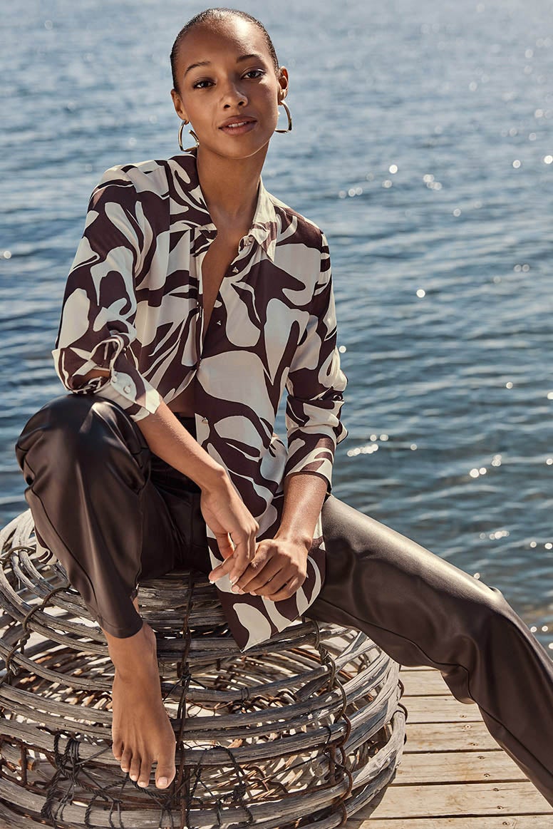 model sits on a fish trap in chocolate brown leather pants and a printed shirt, she is barefoot and calm water is seen in the background