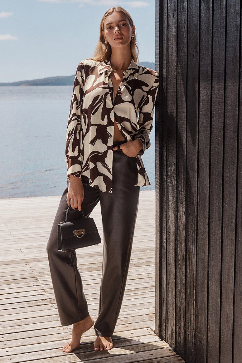 blonde model standing on wharf barefoot in chocolate brown vegan leather pants and a printed brown and white shirt, blue calm water is seen in the background 