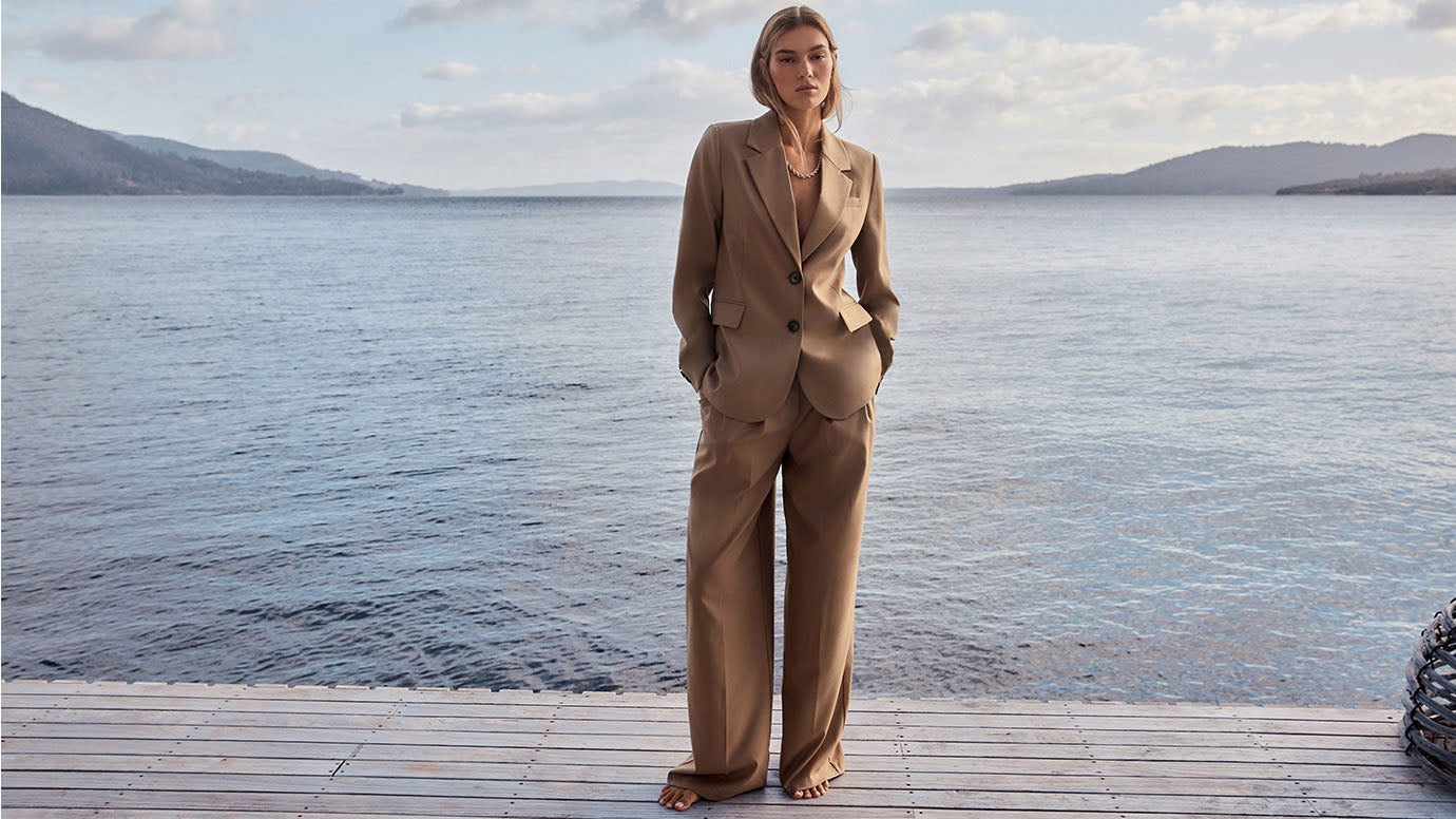 blonde model in caramel suit with no top underneath, she is standing barefoot on a wharf at the waters edge, calm blue waters and sky are seen in the background