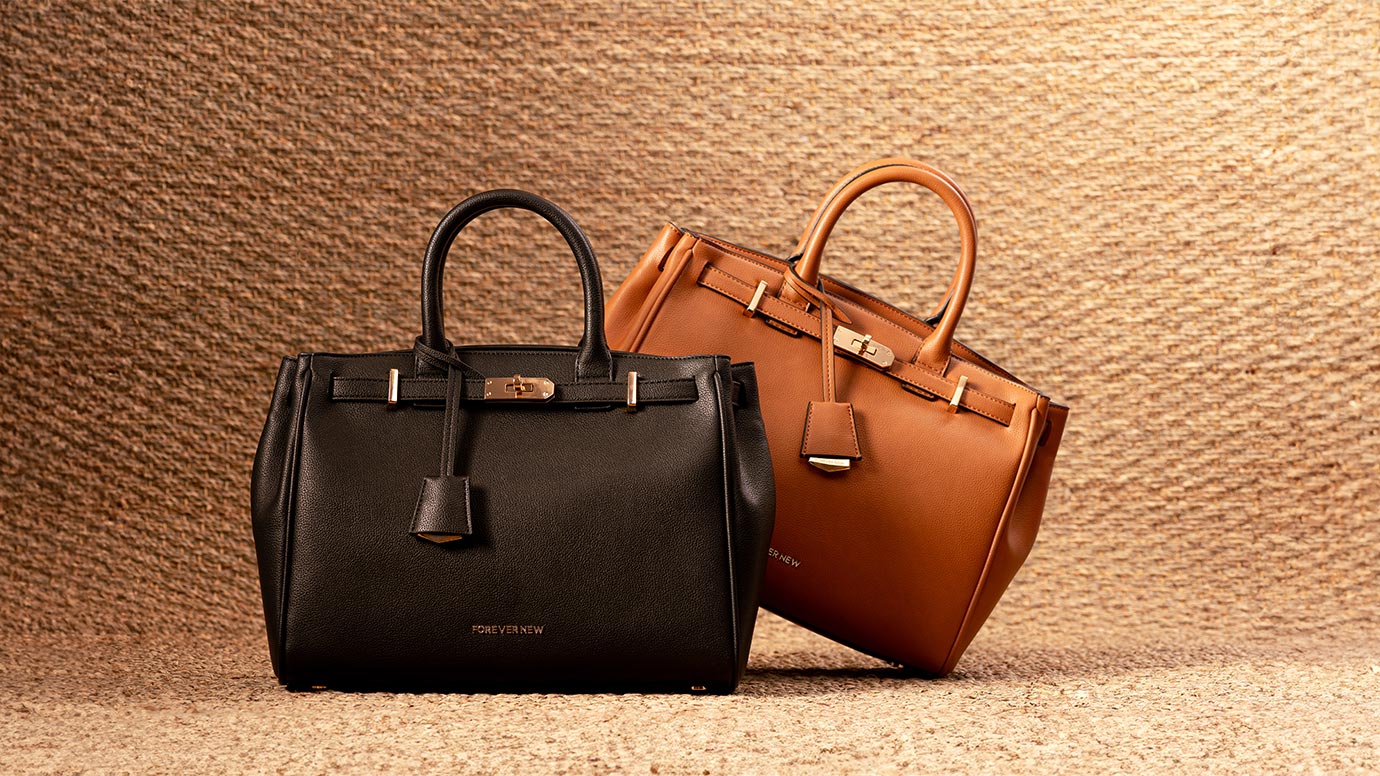 two totes bags that look similar to birkin bags, one black and one caramel tan coloured sitting side by side, the ground and backdrop are warm camel coloured hessian