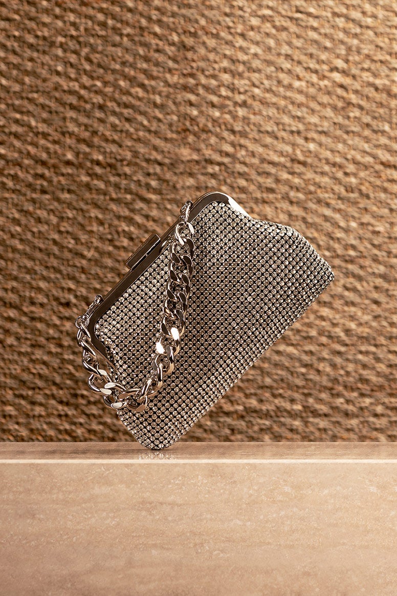 silver glomesh style clutch with a thick short silver link chain handle sitting on a marble looking stone plinth, the background looks like a piece of hessian material