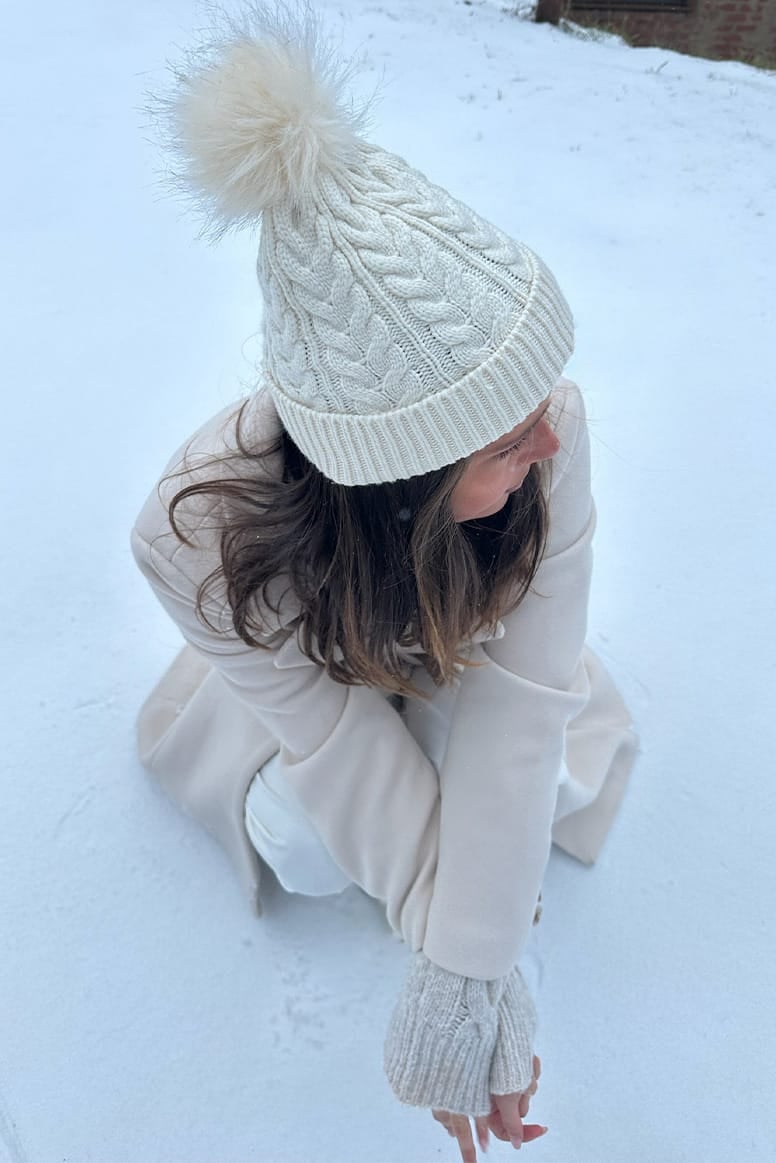 influencer eloise mumme wearing a beige coat, a cream beanie, and white pants with white sneakers kneeling down in the snow