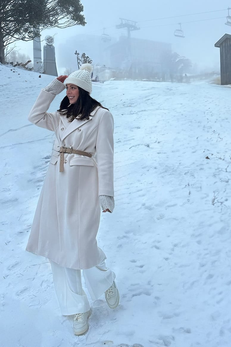 influencer eloise mumme wearing a long beige coat with a brown belt over it white pants white sneakers and a cream beanie she is standing in the snow looking over to the left, you can see the snow resort and ski lift in the background