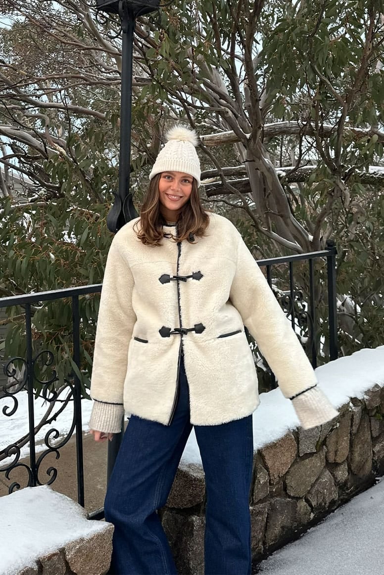 influencer eloise mumme wearing a soft faux fur jacket in cream with black tipping and black toggles down the front, she is wearing dark blue denim and a beanie, behind her is a stone and cast iron fence snow and lots of trees