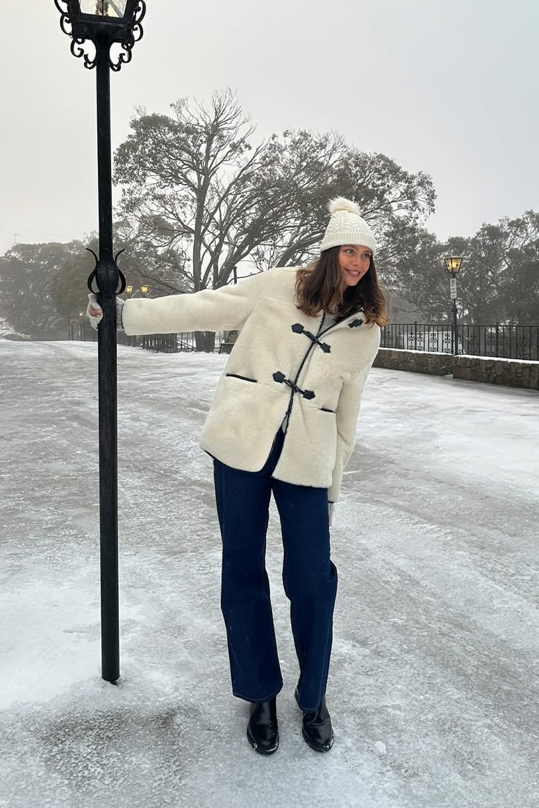influencer eloise mumme wearing faux fur jacket with black toggles a cream pom pom beanie and dark jeans with black boots, she is standing on a snowy road posing holding onto a black vintage street lamp post