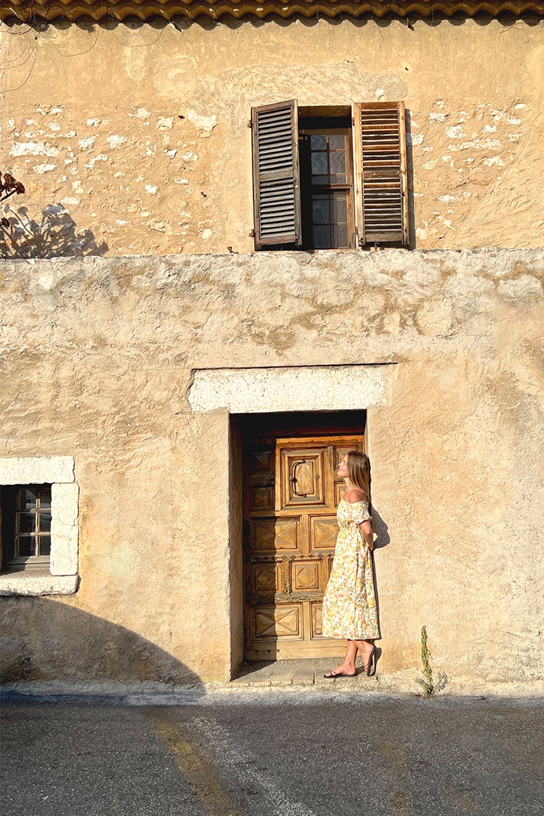 lucy brownless standing in a doorway of an old sandstone building with shutters in a floral bardot sun dress