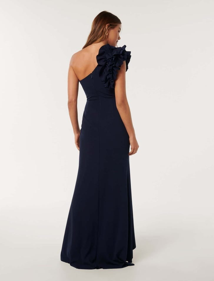 model posing with her back to the camera, she is wearing a a navy maxi dress with a one shoulder design with large ruffle detail