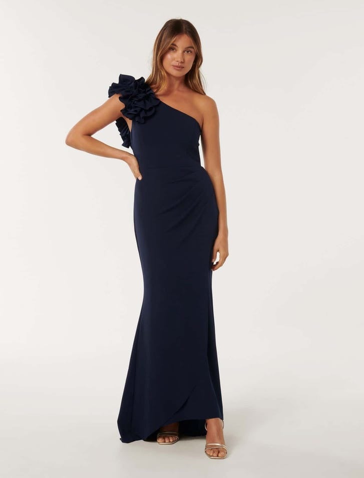 model wearing a one shoulder navy maxi dress with a large ruffle shoulder detail, posing with one hand on hip, she is wearing heels