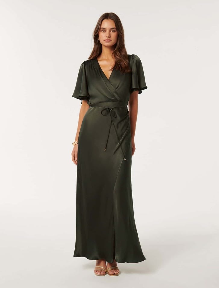 brunette model is wearing a flutter sleeve wrap style dress in dark olive satin, she is wearing strappy gold heels and her arms hung down by her side 