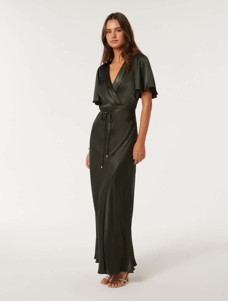 brunette model is wearing a flutter sleeve wrap style dress in dark olive satin, she is wearing strappy gold heels, she is angled toward the camera but standing almost in profile